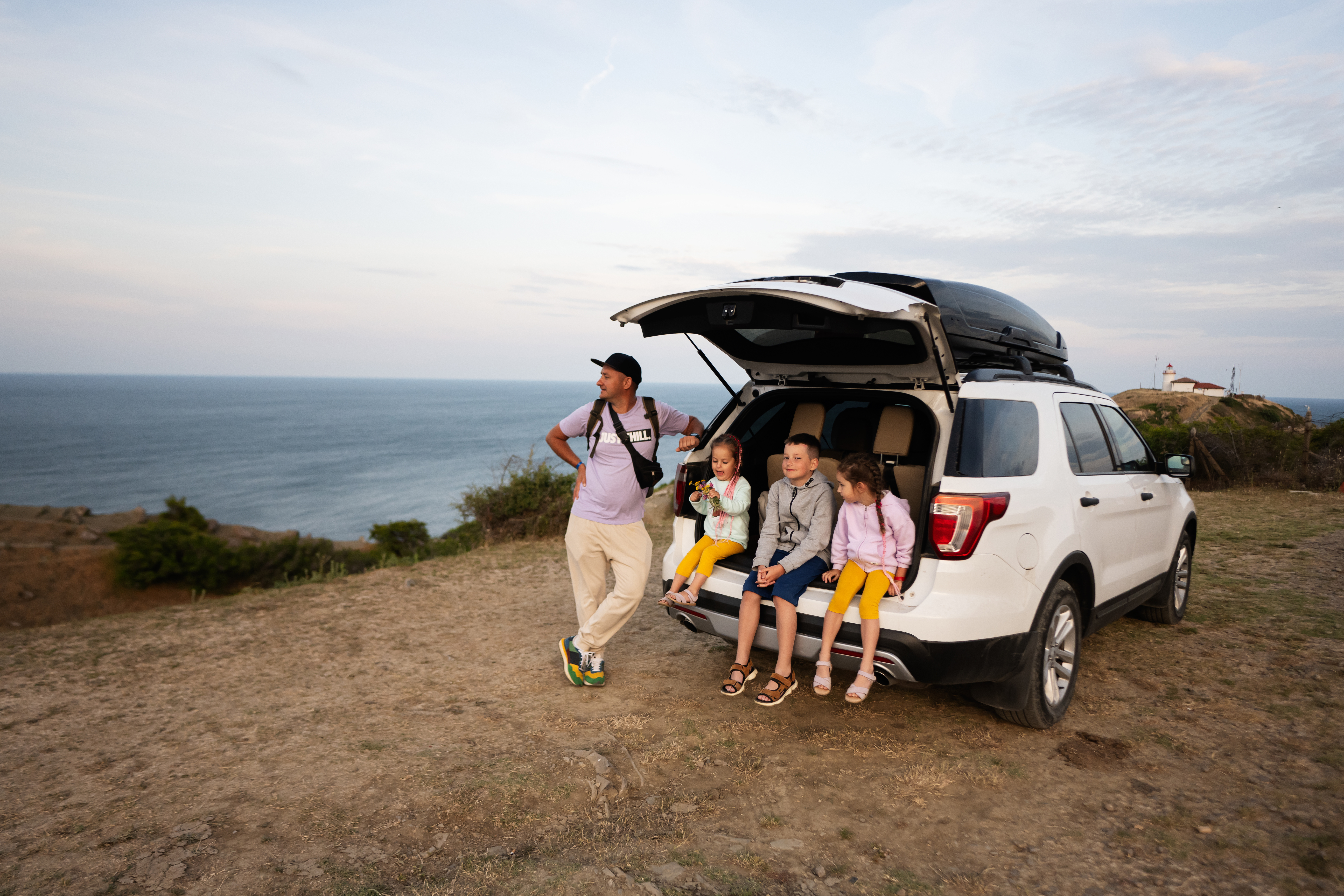 Family sitting on the back of a white suv at the beach against lighthouse. Cape Emine, Black sea coast, Bulgaria.